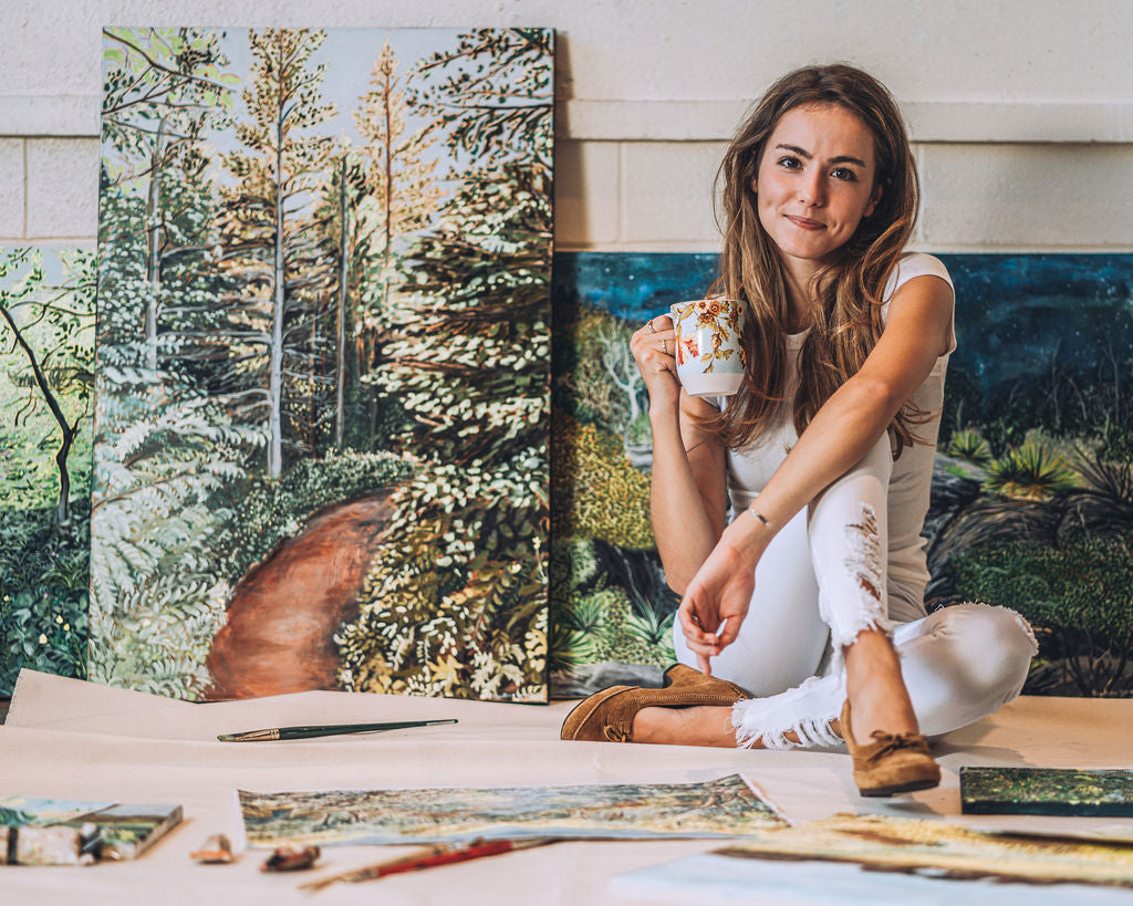 Artist AnnMarie Young sitting in front of her large landscape paintings, depicting Alaska and Texas. 